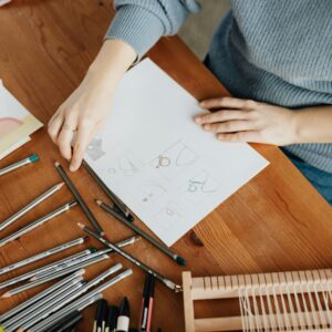 Top view of crop unrecognizable female designer in eyewear sitting at table in front of stationery and wooden weaving loom machine with sheet of paper with drafts